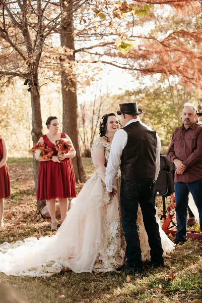 A bride and groom standing face-to-face during an outdoor wedding ceremony in a wooded area, with a bridesmaid and officiant visible in the background.