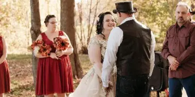 A bride and groom standing face-to-face during an outdoor wedding ceremony in a wooded area, with a bridesmaid and officiant visible in the background.