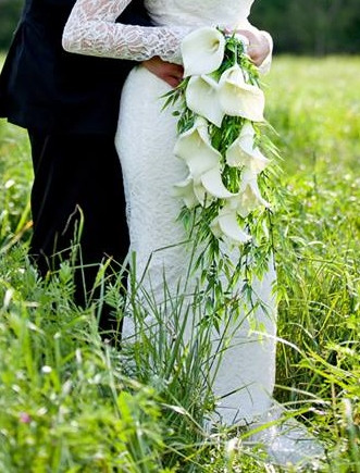 Bride in a white lace gown holding a cascading white calla lily bouquet in a grassy field