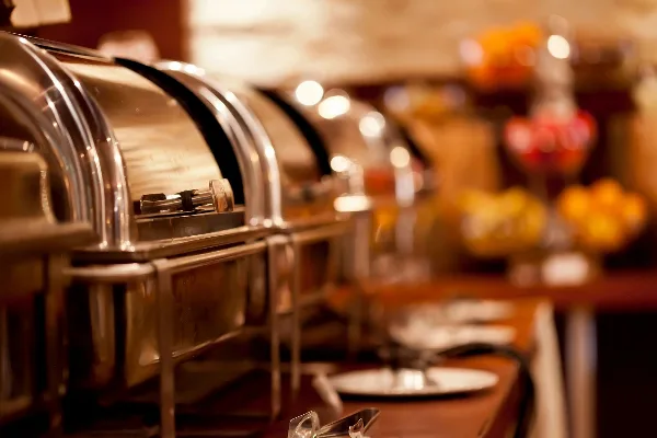 A close-up view of a catering buffet line featuring stainless steel chafing dishes and plates on a table.