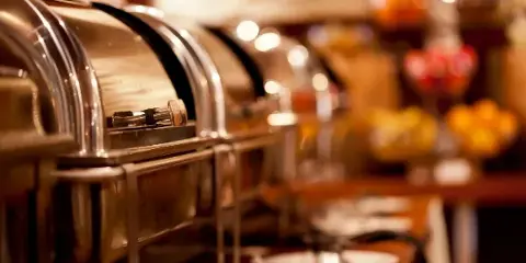 A close-up view of a catering buffet line featuring stainless steel chafing dishes and plates on a table.