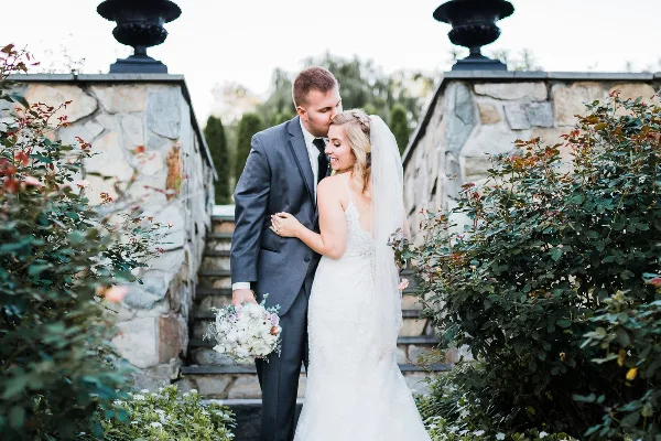 A bride and groom standing together in a garden setting, with the groom kissing the bride's forehead.
