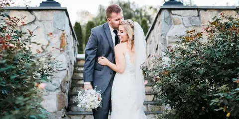 A bride and groom standing together in a garden setting, with the groom kissing the bride's forehead.