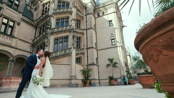 A cinematic wide shot of a bride and groom kissing in front of a large, ornate stone building with terracotta planters.