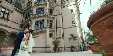 A cinematic wide shot of a bride and groom kissing in front of a large, ornate stone building with terracotta planters.