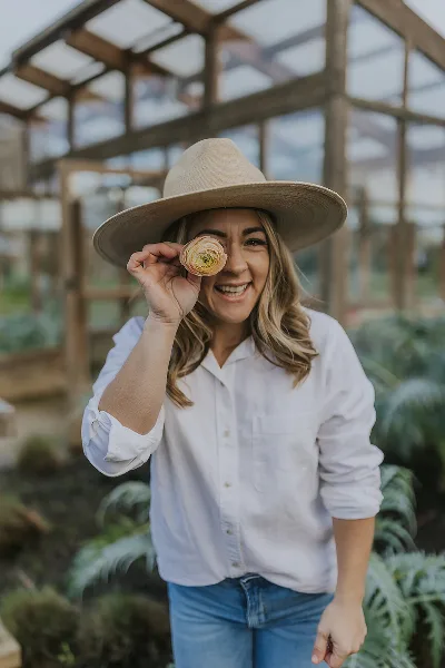A woman in a straw hat and white shirt stands in a greenhouse holding a single flower up to her eye.