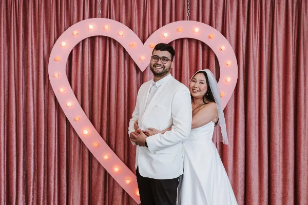 A bride and groom posing together in a studio in front of a large, illuminated heart-shaped prop.