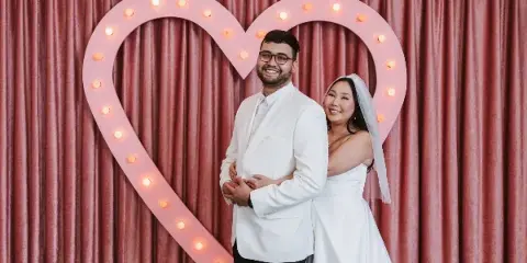 A bride and groom posing together in a studio in front of a large, illuminated heart-shaped prop.