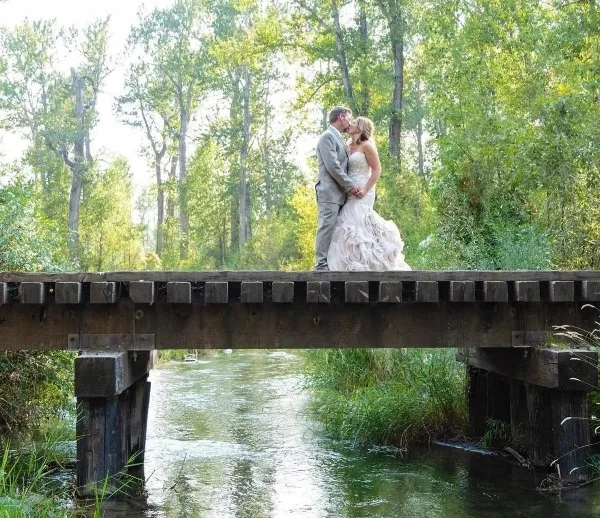 A bride and groom kissing on a wooden railroad bridge over a creek surrounded by trees.
