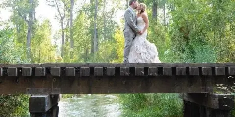 A bride and groom kissing on a wooden railroad bridge over a creek surrounded by trees.