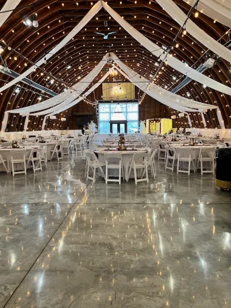 Interior of a wedding venue barn featuring a high wooden ceiling, string lights, and round tables set for a reception.