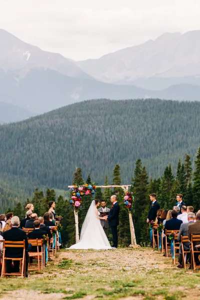 A wedding ceremony taking place outdoors with a bride, groom, and officiant at an altar, set against a backdrop of mountains and pine trees.