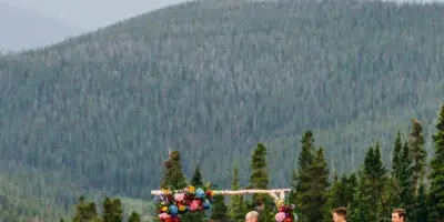A wedding ceremony taking place outdoors with a bride, groom, and officiant at an altar, set against a backdrop of mountains and pine trees.