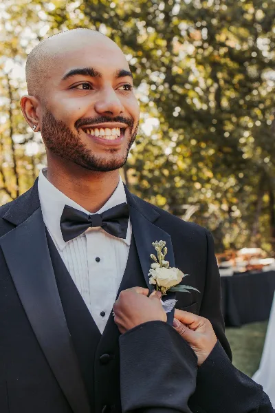 A smiling groom in a black tuxedo with a white rose boutonniere at an outdoor wedding.