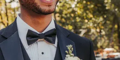 A smiling groom in a black tuxedo with a white rose boutonniere at an outdoor wedding.