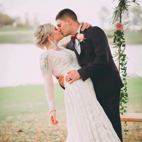 A bride and groom sharing a kiss outdoors near a floral arch and a body of water.