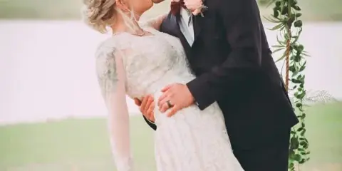 A bride and groom sharing a kiss outdoors near a floral arch and a body of water.