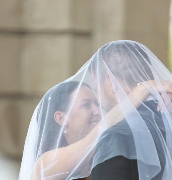 A bride and groom embracing under a white wedding veil in a candid portrait.