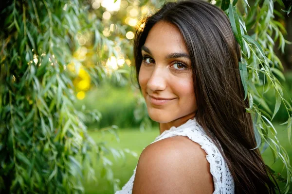 A professional outdoor portrait of a young woman with long dark hair, smiling and looking over her shoulder, set against a background of green willow branches and soft sunlight.