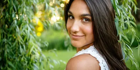 A professional outdoor portrait of a young woman with long dark hair, smiling and looking over her shoulder, set against a background of green willow branches and soft sunlight.