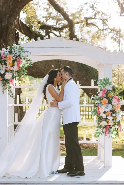 A bride and groom sharing a kiss at a white wooden ceremony arch decorated with floral arrangements on the grounds of Chatsworth Plantation.