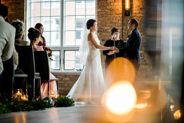 A bride and groom holding hands during a wedding ceremony with an officiant at a microphone in a brick-walled venue.