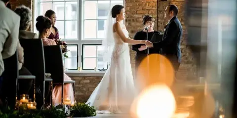 A bride and groom holding hands during a wedding ceremony with an officiant at a microphone in a brick-walled venue.