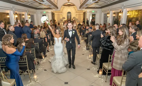 A bride and groom walking down the aisle at a wedding reception surrounded by seated guests.