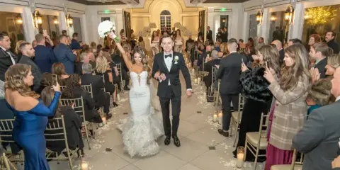 A bride and groom walking down the aisle at a wedding reception surrounded by seated guests.