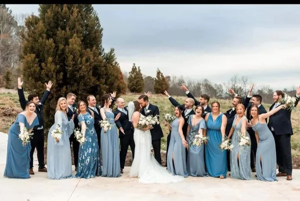 A wedding party including a bride and groom posing outdoors with raised hands.