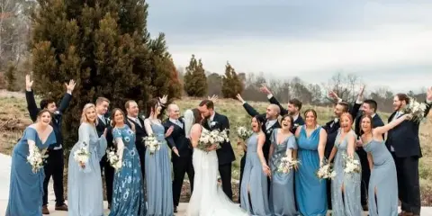 A wedding party including a bride and groom posing outdoors with raised hands.