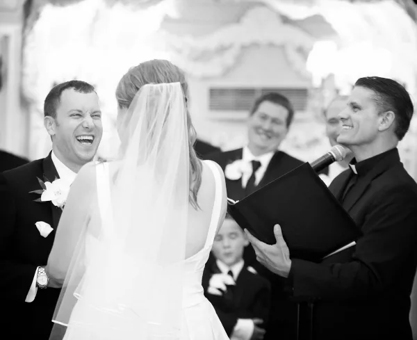 A wedding officiant in clerical attire speaks into a microphone while facing a bride and groom during a ceremony.
