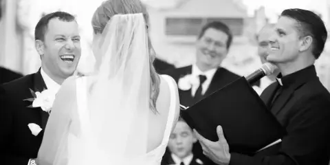 A wedding officiant in clerical attire speaks into a microphone while facing a bride and groom during a ceremony.