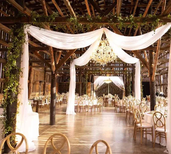 A rustic wooden wedding venue interior featuring white draped fabric, a chandelier, and set dining tables with gold chairs.