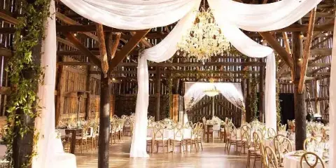 A rustic wooden wedding venue interior featuring white draped fabric, a chandelier, and set dining tables with gold chairs.