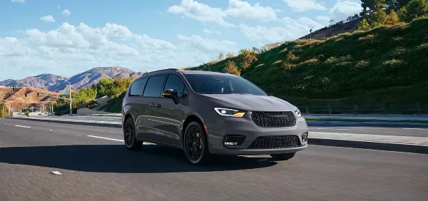 A grey Chrysler Pacifica minivan driving on a paved road with a grassy hill and mountains in the background.