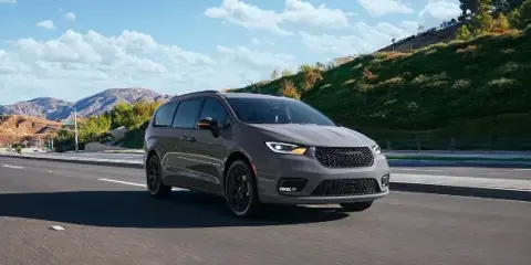 A grey Chrysler Pacifica minivan driving on a paved road with a grassy hill and mountains in the background.