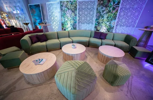 A modern lounge setup featuring a green sectional sofa, matching ottomans, and round coffee tables arranged in an event space.