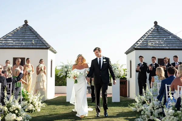 A bride and groom walking down the aisle at an outdoor wedding ceremony with guests and floral arrangements.
