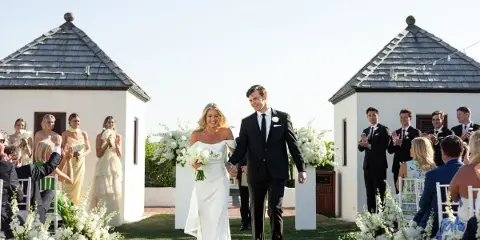 A bride and groom walking down the aisle at an outdoor wedding ceremony with guests and floral arrangements.