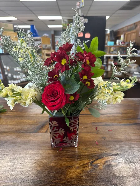 A fresh floral arrangement with red roses and chrysanthemums in a red patterned glass vase on a wooden table.