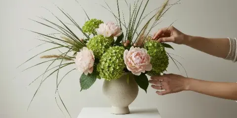 A pair of hands arranging a floral design featuring pink peonies, green hydrangeas, and tall grasses in a cream-colored vase.