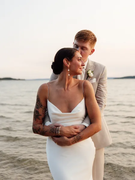 A bride and groom embracing on a beach at sunset, with the groom kissing the bride's forehead.
