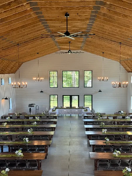 Interior view of a large barn wedding venue with high wooden ceilings, modern chandeliers, and long tables set for a reception.
