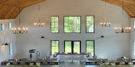 Interior view of a large barn wedding venue with high wooden ceilings, modern chandeliers, and long tables set for a reception.
