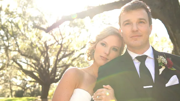 A bride in a white strapless gown and a groom in a black suit with a boutonniere posing together outdoors under trees.