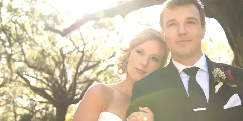 A bride in a white strapless gown and a groom in a black suit with a boutonniere posing together outdoors under trees.