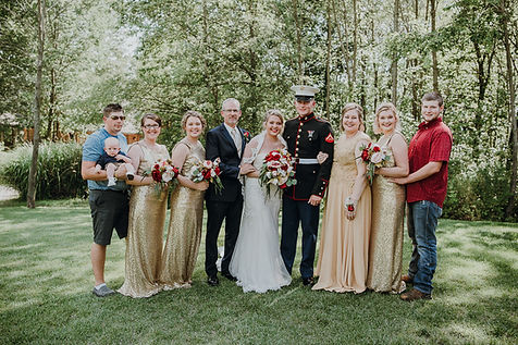 A wedding party consisting of a bride, groom, and attendants posing for a group photo on a grassy lawn in a wooded area.