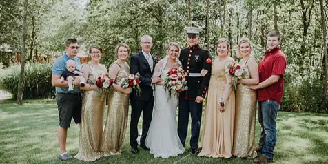 A wedding party consisting of a bride, groom, and attendants posing for a group photo on a grassy lawn in a wooded area.