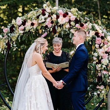 A wedding officiant in a black dress stands between a bride and groom, holding a book and officiating their ceremony under a floral arch.
